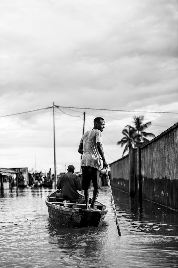 about-us Black and white photo of two men navigating a flooded rural village by boat.