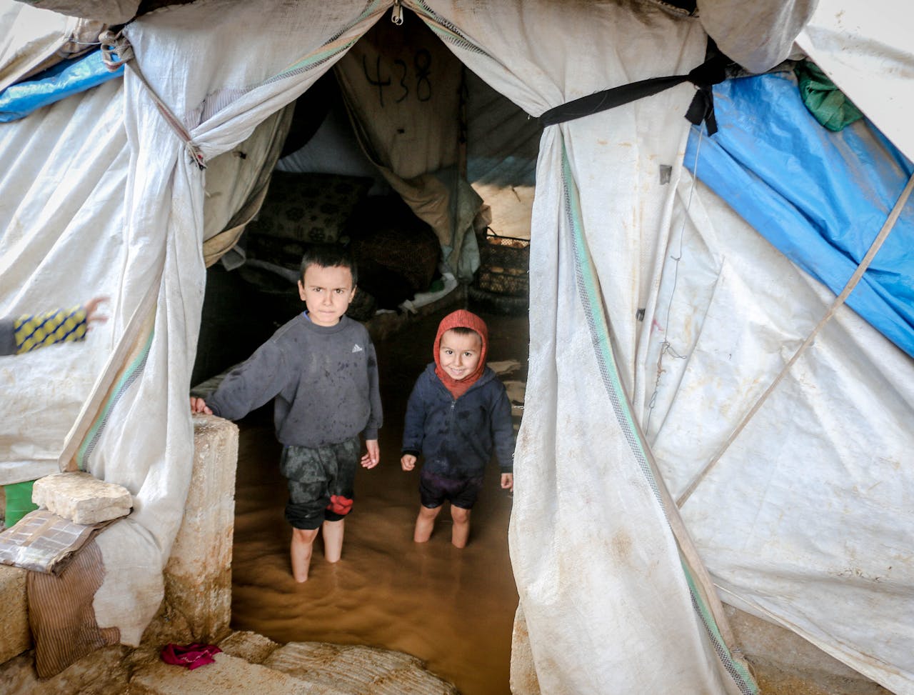 services-03 Two young children stand in a flooded tent in Idlib, Syria, highlighting the hardships in refugee camps.