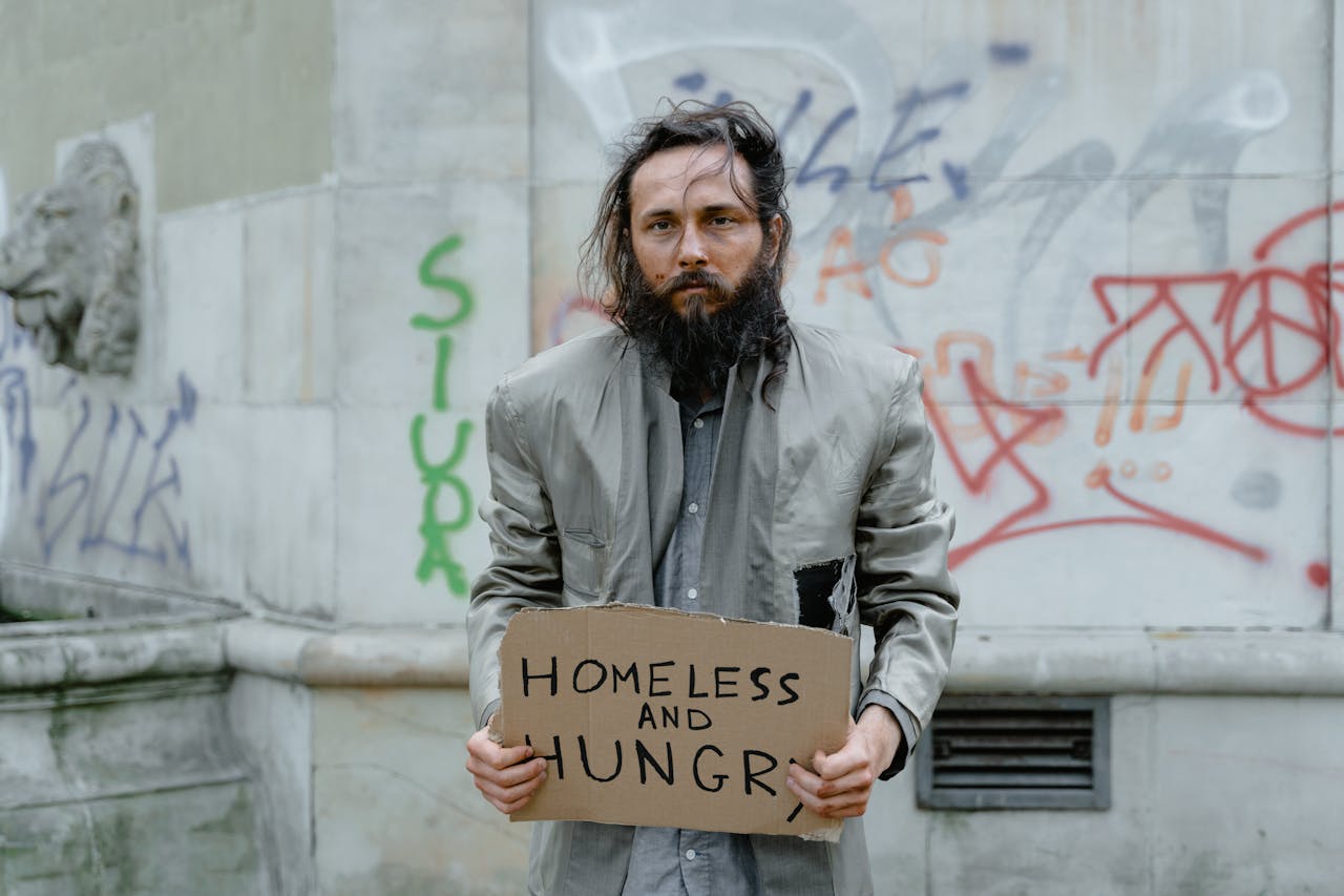 about-us Homeless man standing on the street holding a sign, with graffiti in the background.